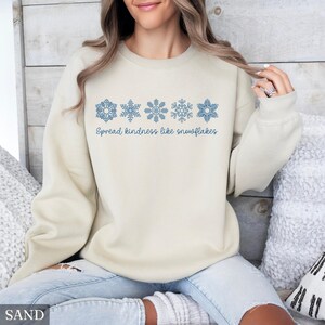 May include: A sand-colored sweatshirt with blue snowflake designs and the text "Spread kindness like snowflakes." The sweatshirt is a neutral color and features a crew neck. The design is a winter theme.