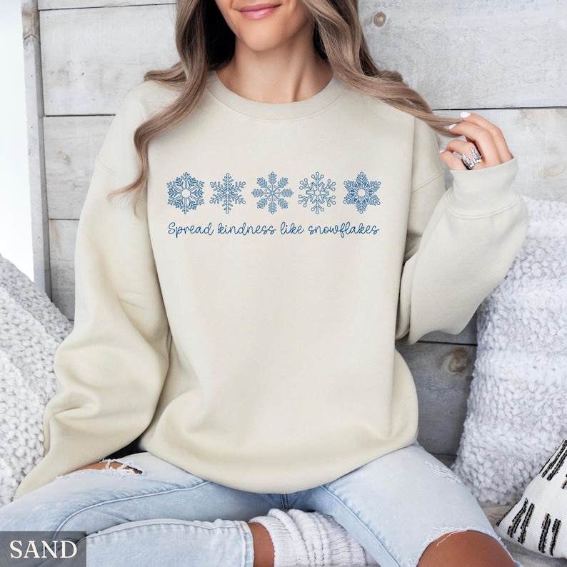 May include: A sand-colored sweatshirt with blue snowflake designs and the text "Spread kindness like snowflakes." The sweatshirt is a neutral color and features a crew neck. The design is a winter theme.