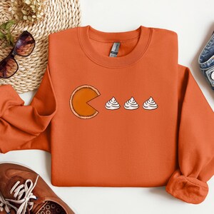 Pode incluir: Camisola laranja com um gr&aacute;fico de uma fatia de torta de ab&oacute;bora em forma de Pac-Man comendo tr&ecirc;s mini tortas de ab&oacute;bora.