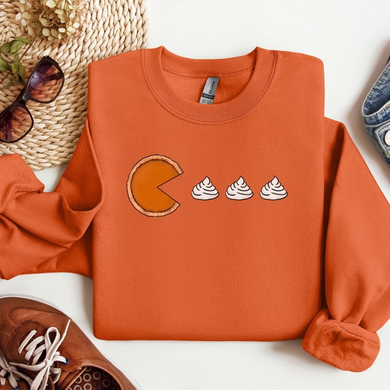 Pode incluir: Camisola laranja com um gr&aacute;fico de uma fatia de torta de ab&oacute;bora em forma de Pac-Man comendo tr&ecirc;s mini tortas de ab&oacute;bora.