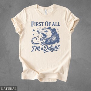 May include: Cream-colored t-shirt with rolled-up sleeves. The front features a blue graphic of an opossum with an open mouth, accompanied by the text "FIRST OF ALL I'M a Delight" in blue. The shirt is made of a soft material.