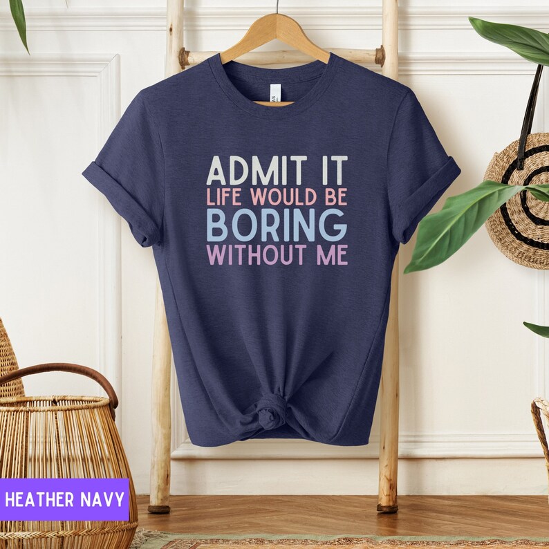 May include: Heather navy t-shirt with the text "ADMIT IT LIFE WOULD BE BORING WITHOUT ME" in white, light blue, and pink. The t-shirt is knotted at the waist and hangs on a wooden hanger.