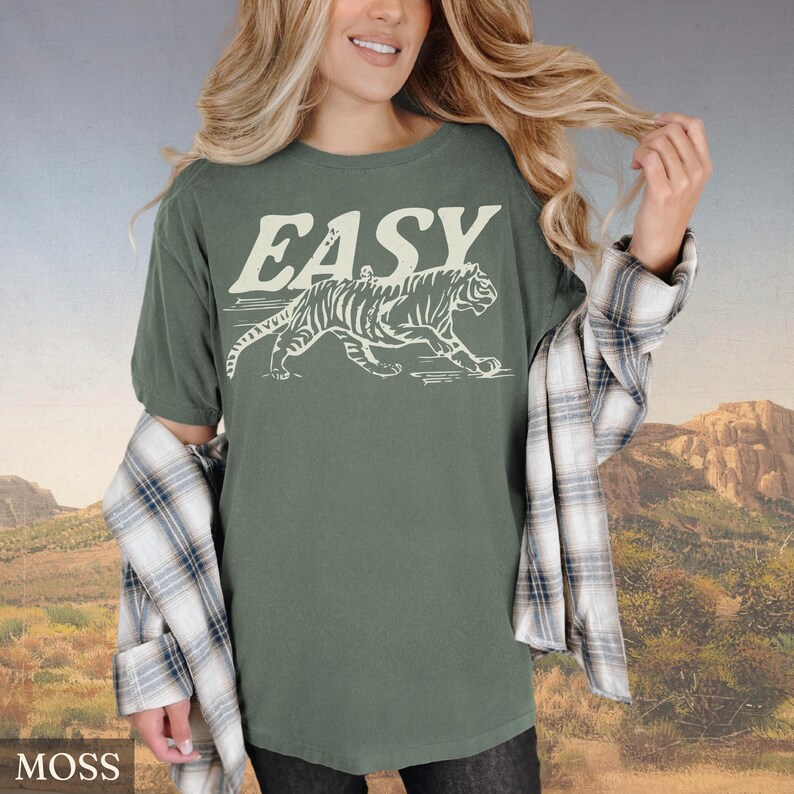Op de afbeelding: Mosgroen t-shirt met het woord "EASY" in een vintage lettertype en een witte tijgerafbeelding. Het shirt heeft een relaxte pasvorm en korte mouwen. Het model draagt een geruit overhemd over het t-shirt.