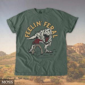 May include: Moss green t-shirt with a graphic of a cowboy roping a Tyrannosaurus Rex. The text "FEELIN' FERAL" is arched above the dinosaur. The shirt is a solid color with a crew neck and short sleeves.