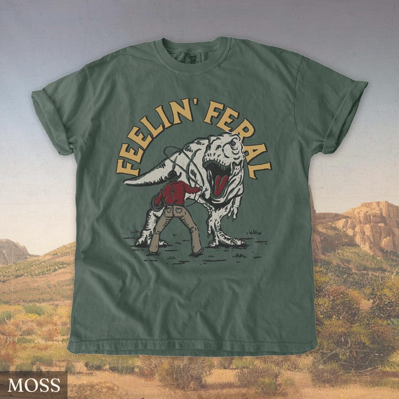 May include: Moss green t-shirt with a graphic of a cowboy roping a Tyrannosaurus Rex. The text "FEELIN' FERAL" is arched above the dinosaur. The shirt is a solid color with a crew neck and short sleeves.