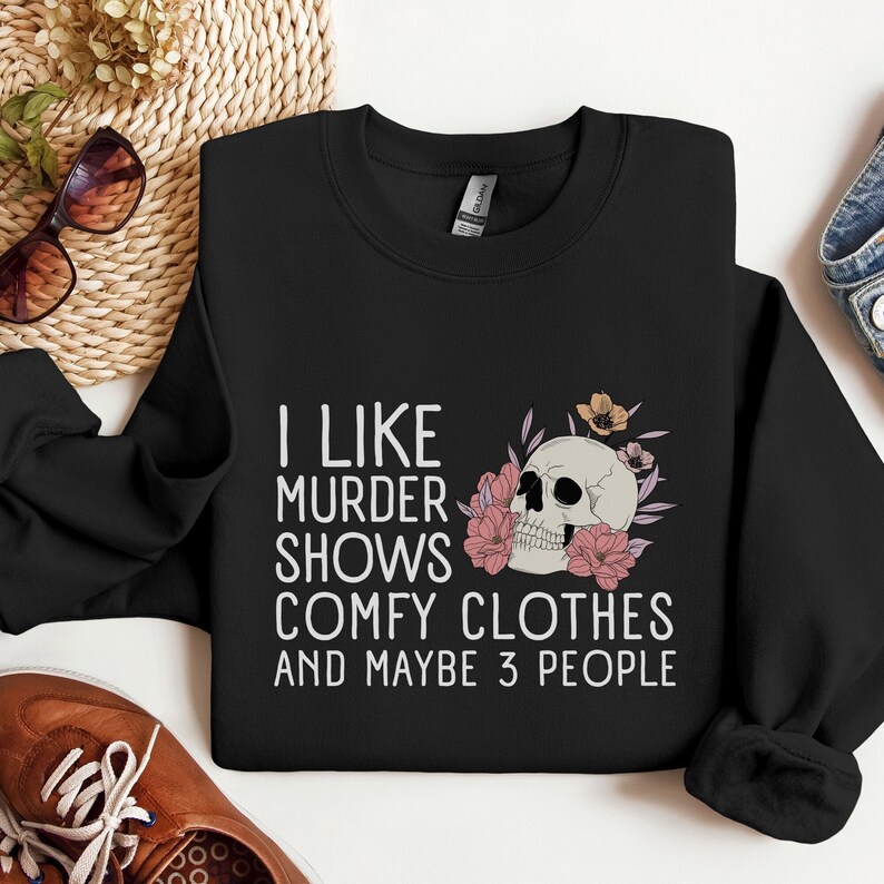 Op de afbeelding: Zwart sweatshirt met een witte afbeelding die zegt "Ik hou van moordzaken, comfortabele kleding en misschien 3 mensen". De afbeelding bevat ook een schedel met bloemen.