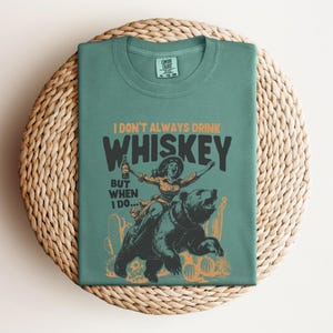 May include: A teal green t-shirt with a graphic of a woman riding a bear. The text on the shirt reads "I don't always drink whiskey, but when I do..."