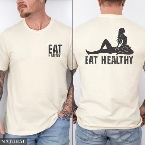 May include: Natural-colored t-shirt with the text "EAT HEALTHY" printed on the front and back. The back of the shirt features a silhouette of a woman. The shirt is made of a soft material.