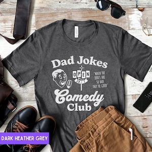 May include: Dark heather grey t-shirt with a white graphic of a retro comedy club sign that says "Dad Jokes Comedy Club" and "When the jokes are so bad they're good".