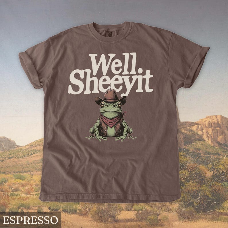 May include: Espresso-colored t-shirt featuring a cartoon frog wearing a cowboy hat, vest, and red bandana. The text "Well. Sheeeyit" is printed above the frog. The shirt is displayed against a desert landscape backdrop.