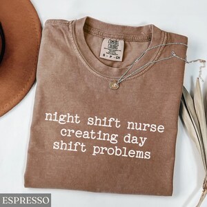 May include: Espresso-colored t-shirt with the text "night shift nurse creating day shift problems" in white. A silver necklace with a pendant is draped on the shirt. The shirt is a Comfort Colors brand.