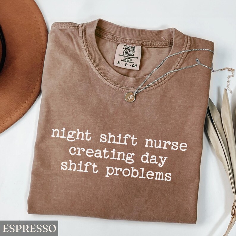 May include: Espresso-colored t-shirt with the text "night shift nurse creating day shift problems" in white. A silver necklace with a pendant is draped on the shirt. The shirt is a Comfort Colors brand.