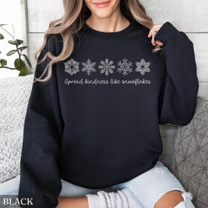 May include: Black sweatshirt with white snowflake designs across the chest. The text "Spread kindness like snowflakes" is printed below the snowflakes. The word "BLACK" is printed at the bottom left corner of the image.