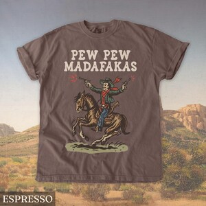 Puede incluir: Camiseta color espresso con el texto "PEW PEW MADAF*KAS" en blanco. El gr&aacute;fico muestra un vaquero a caballo disparando pistolas. El vaquero lleva una chaqueta verde, una bufanda roja y un sombrero de vaquero.