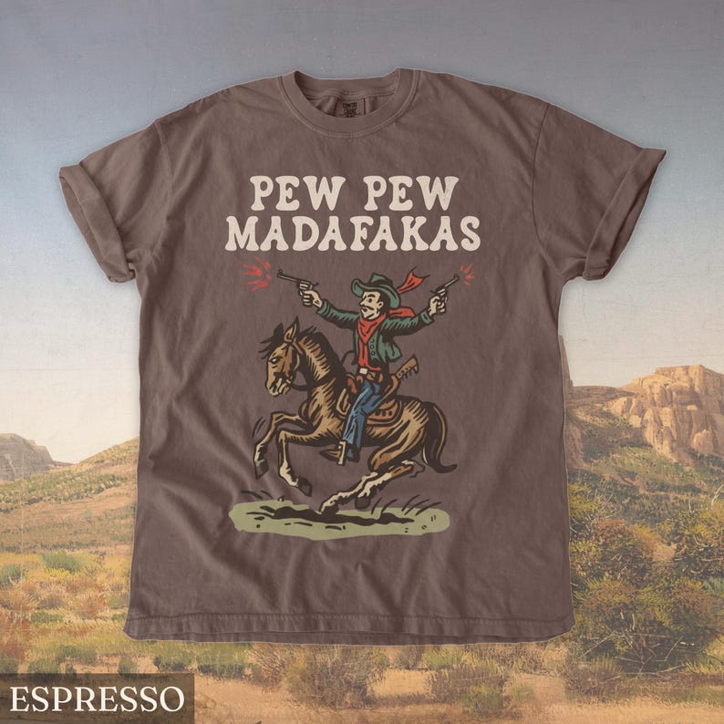 Puede incluir: Camiseta color espresso con el texto "PEW PEW MADAF*KAS" en blanco. El gr&aacute;fico muestra un vaquero a caballo disparando pistolas. El vaquero lleva una chaqueta verde, una bufanda roja y un sombrero de vaquero.