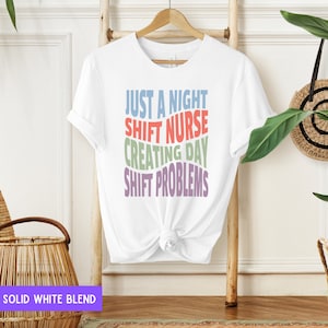 May include: White t-shirt with the text "Just a night shift nurse creating day shift problems" in colourful, wavy letters.