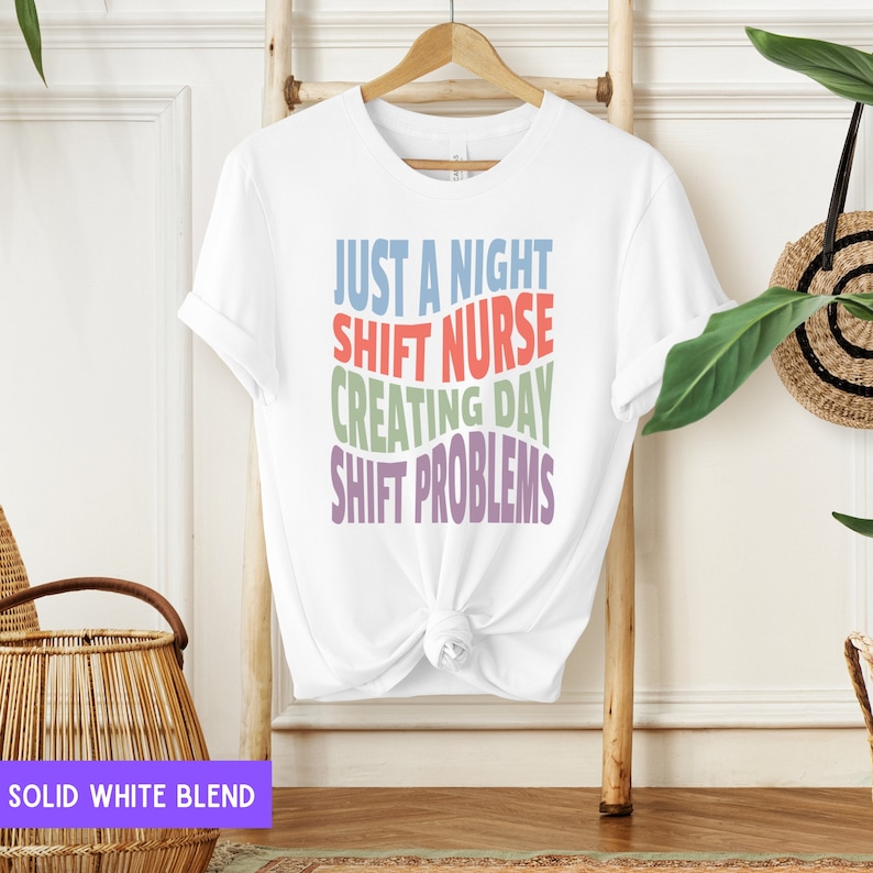 May include: White t-shirt with the text "Just a night shift nurse creating day shift problems" in colourful, wavy letters.