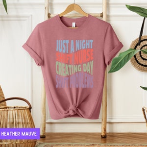 May include: A heather mauve t-shirt with the text "Just a night shift nurse creating day shift problems" in colourful, stacked text.