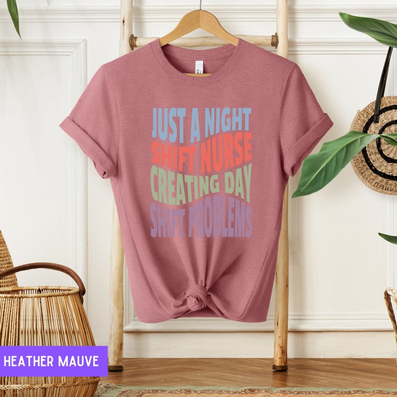 May include: A heather mauve t-shirt with the text "Just a night shift nurse creating day shift problems" in colourful, stacked text.