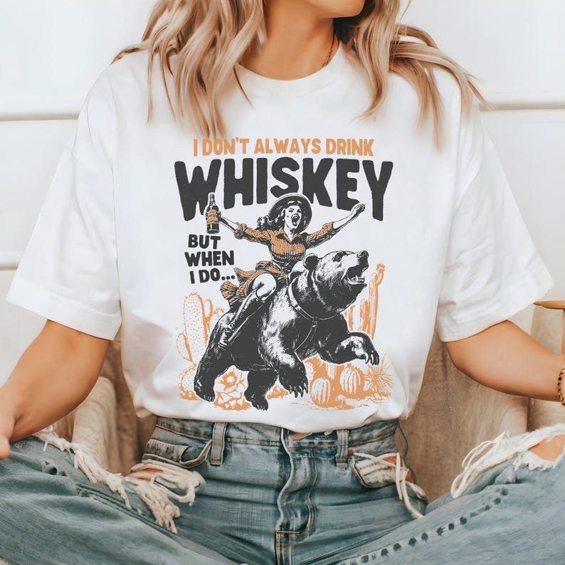 May include: White t-shirt with a graphic of a woman riding a bear and holding a bottle of whiskey. The text on the shirt reads "I don't always drink whiskey but when I do..."