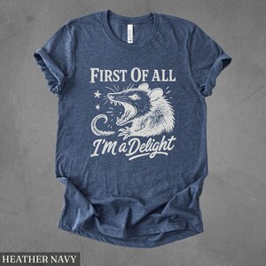 May include: Heather navy blue t-shirt with a white graphic of a possum with an open mouth and the text "FIRST OF ALL I'M A DELIGHT". The shirt has short sleeves and a crew neck.