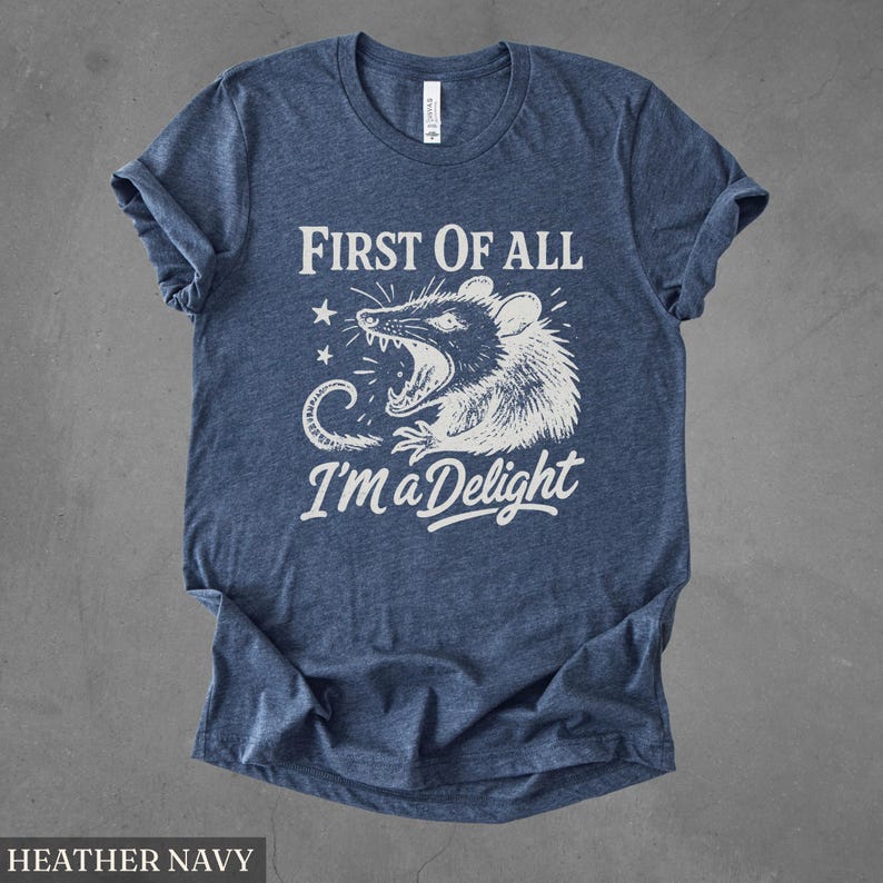 May include: Heather navy blue t-shirt with a white graphic of a possum with an open mouth and the text "FIRST OF ALL I'M A DELIGHT". The shirt has short sleeves and a crew neck.