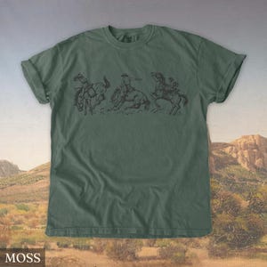 Puede incluir: Camiseta verde musgo con un gr&aacute;fico negro de tres vaqueros montando caballos salvajes. El dise&ntilde;o representa una escena de rodeo con jinetes tratando de mantenerse sobre sus caballos. La camiseta tiene cuello redondo y mangas cortas.