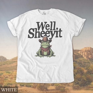 May include: White t-shirt with the text "Well. Sheeeyit" above a cartoon frog wearing a cowboy hat, vest, and red bandana. The frog is green with brown and tan accents. The shirt has rolled-up sleeves.