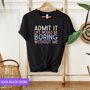 May include: Black t-shirt with the text "ADMIT IT LIFE WOULD BE BORING WITHOUT ME" in white, pink, and blue. The shirt is hanging on a wooden hanger against a light background.