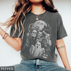 May include: A charcoal gray short-sleeve t-shirt featuring a graphic print of classic horror movie monsters including Frankenstein, a mummy, a vampire, a bride, a werewolf, and a ghost, set against a full moon and a dark castle background.