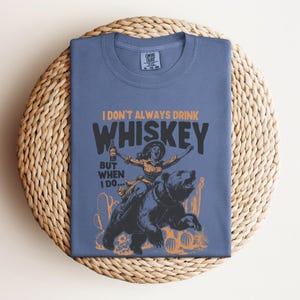 May include: A blue t-shirt with a graphic of a woman riding a bear. The text on the shirt reads "I don't always drink whiskey, but when I do..."