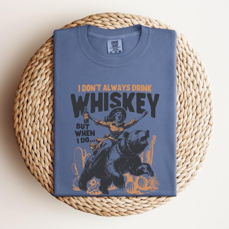May include: A blue t-shirt with a graphic of a woman riding a bear. The text on the shirt reads "I don't always drink whiskey, but when I do..."