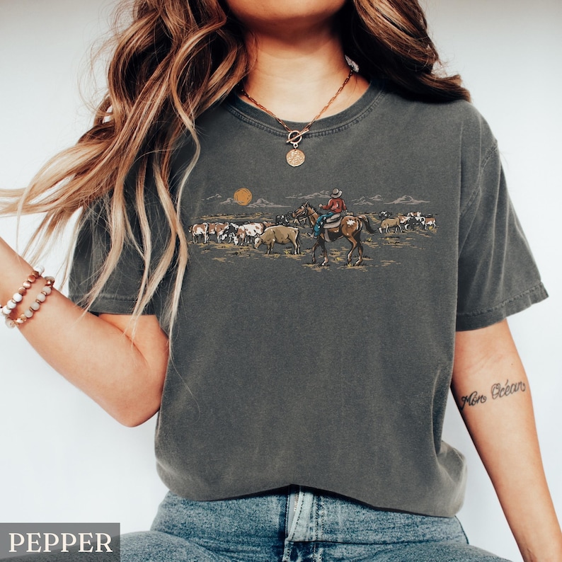May include: Pepper-colored t-shirt featuring a Western-themed graphic. The design depicts a cowboy on horseback herding cattle under a setting sun. The shirt is a relaxed fit, perfect for casual wear.