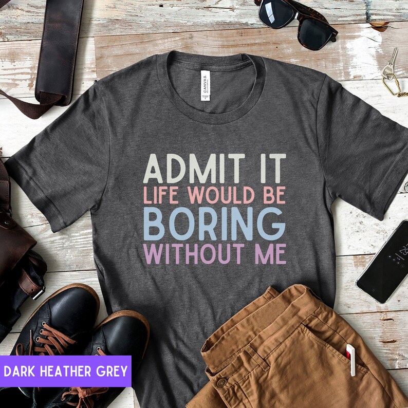 May include: Dark heather grey t-shirt with the text "ADMIT IT LIFE WOULD BE BORING WITHOUT ME" in white, pink, and blue. The shirt is displayed with brown pants, black shoes, and accessories.