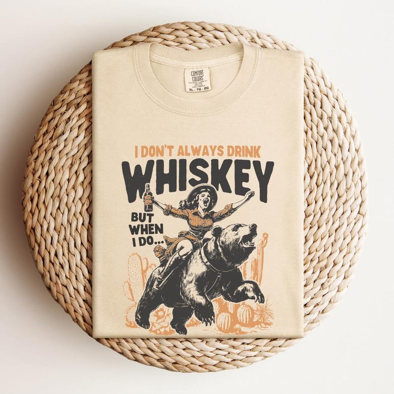 May include: A beige t-shirt with a graphic print of a woman riding a bear and holding a bottle of whiskey. The text on the shirt reads "I don't always drink whiskey, but when I do..."