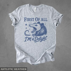 May include: Athletic heather gray t-shirt with a blue graphic of an opossum with the text "First of All I'm a Delight". The shirt has rolled-up sleeves and a crew neck.