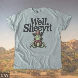 May include: Light blue t-shirt with the text "Well. Sheeeyit" above a cartoon frog wearing a cowboy hat, vest, and bandana. The frog is green with brown and red accents. The shirt has rolled-up sleeves.