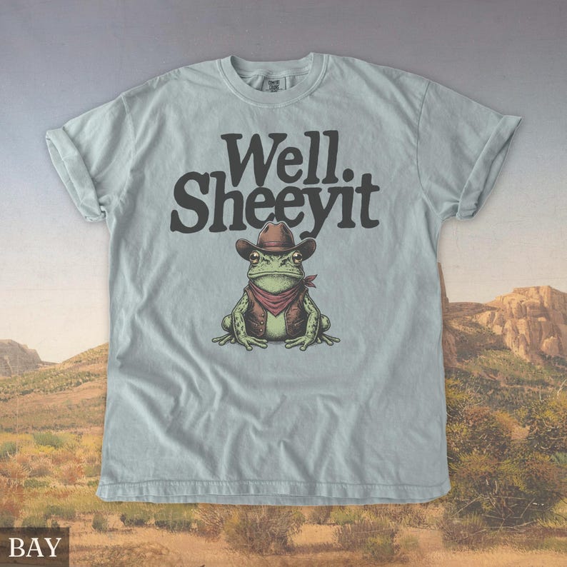 May include: Light blue t-shirt with the text "Well. Sheeeyit" above a cartoon frog wearing a cowboy hat, vest, and bandana. The frog is green with brown and red accents. The shirt has rolled-up sleeves.