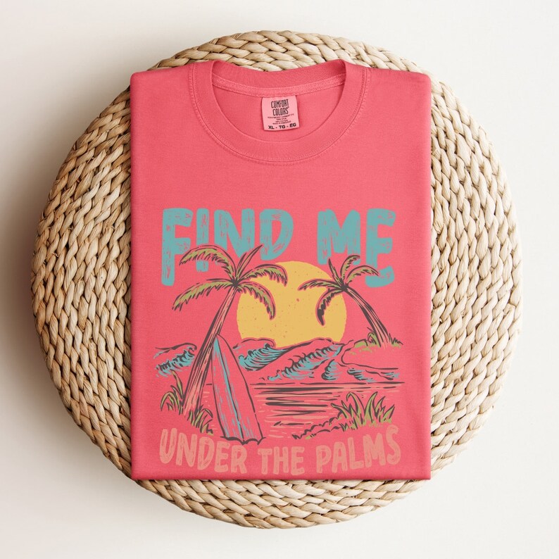 May include: A coral pink t-shirt with a graphic design of palm trees, waves, and a surfboard. The text "FIND ME UNDER THE PALMS" is printed on the shirt.