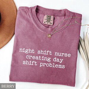 May include: Berry-colored t-shirt with the text "night shift nurse creating day shift problems" in white. A gold necklace with a pendant is draped across the shirt. The shirt is made of a soft material.