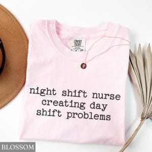 May include: A light pink t-shirt with the text "night shift nurse creating day shift problems" in black. A gold necklace with a small pendant is on the shirt. A brown hat and a dried palm leaf are in the background.