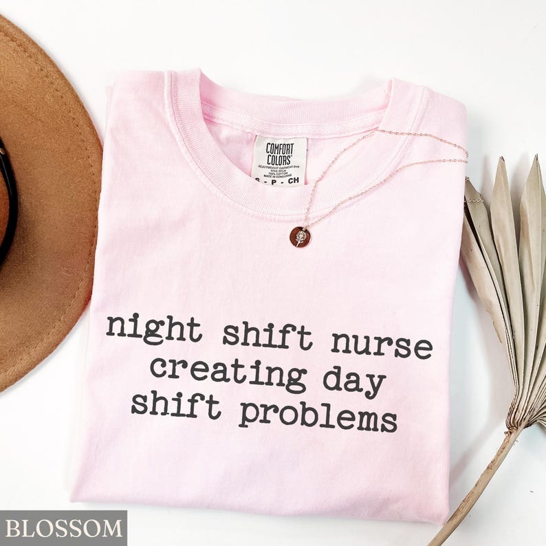 May include: A light pink t-shirt with the text "night shift nurse creating day shift problems" in black. A gold necklace with a small pendant is on the shirt. A brown hat and a dried palm leaf are in the background.