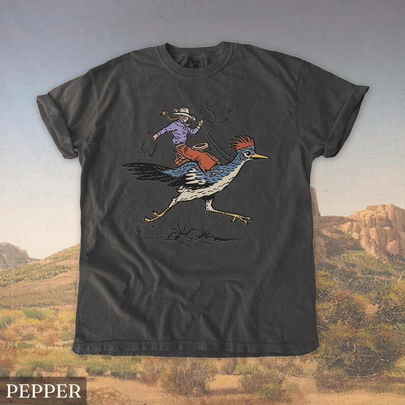 May include: Dark gray t-shirt featuring a cartoon illustration of a cowboy riding a roadrunner. The cowboy wears a purple shirt, red chaps, and a white hat, holding a lasso. The roadrunner is blue, white, and black. The word "PEPPER" is at the bottom.