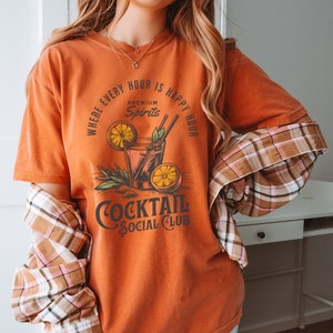 May include: Orange t-shirt with a graphic of a cocktail glass with lemons and the text "Where every hour is happy hour, Premium Spirits, Cocktail Social Club".