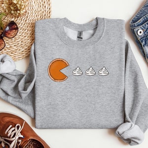 Pode incluir: Uma camisa de moletom cinza com um gr&aacute;fico de uma fatia de torta de ab&oacute;bora comendo tr&ecirc;s colheres de sopa de chantilly. O gr&aacute;fico &eacute; uma par&oacute;dia do cl&aacute;ssico videogame Pac-Man.
