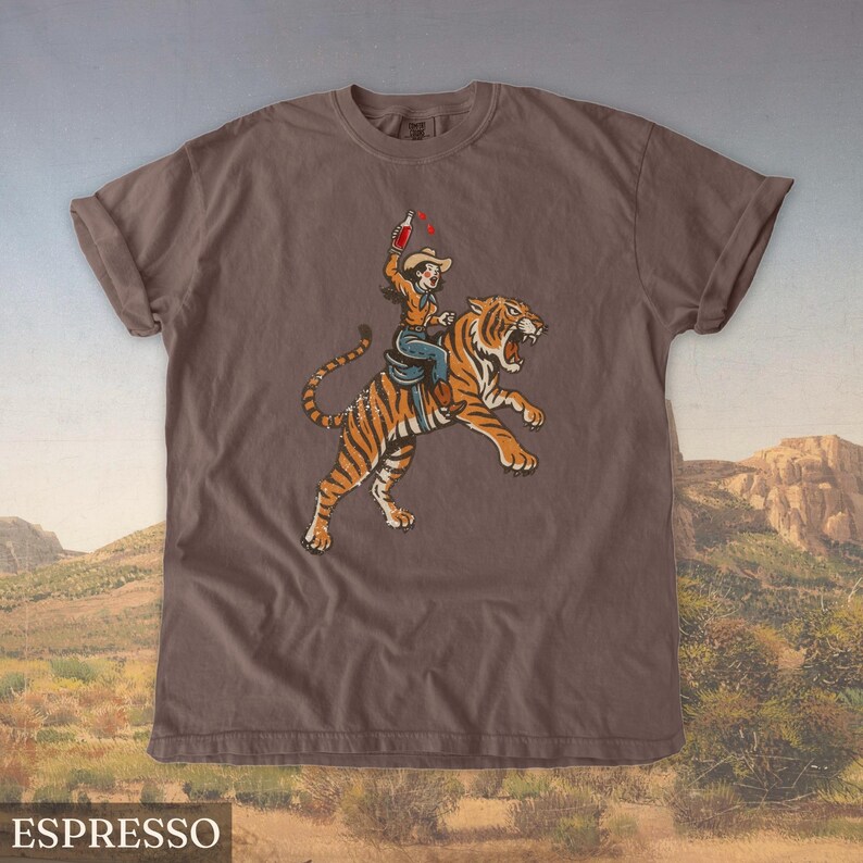 May include: Espresso-colored t-shirt featuring a graphic of a person riding a tiger, holding a bottle. The tiger is orange with black stripes. The person is wearing a cowboy hat and blue jeans. The shirt has rolled-up sleeves.
