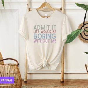 May include: Cream-colored t-shirt with the text "ADMIT IT LIFE WOULD BE BORING WITHOUT ME" in shades of green, blue, and purple. The shirt is hanging on a wooden hanger against a light background.