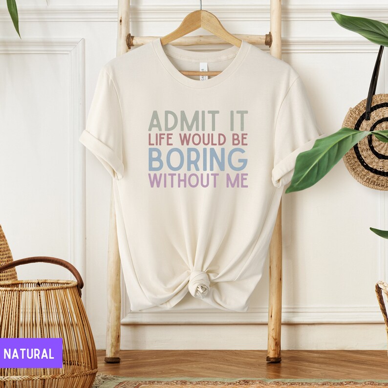 May include: Cream-colored t-shirt with the text "ADMIT IT LIFE WOULD BE BORING WITHOUT ME" in shades of green, blue, and purple. The shirt is hanging on a wooden hanger against a light background.
