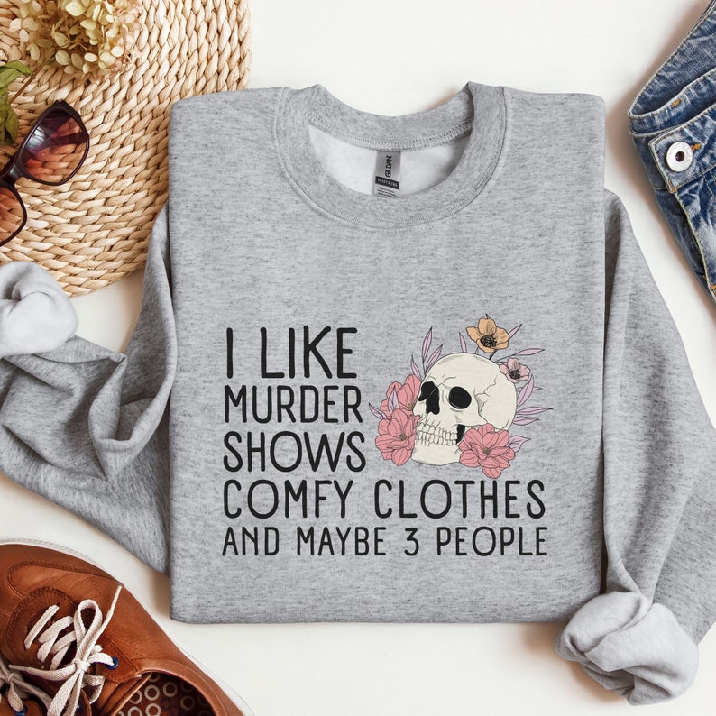 Op de afbeelding: Grijs sweatshirt met een schedel- en bloemenprint en de tekst "Ik hou van moordshows, comfortabele kleding en misschien 3 mensen".