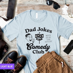 May include: Light blue t-shirt with a black graphic of a man with a microphone and the text "Dad Jokes Comedy Club" with a sign that says "Open 24/7" and the text "When the jokes are so bad they're good."